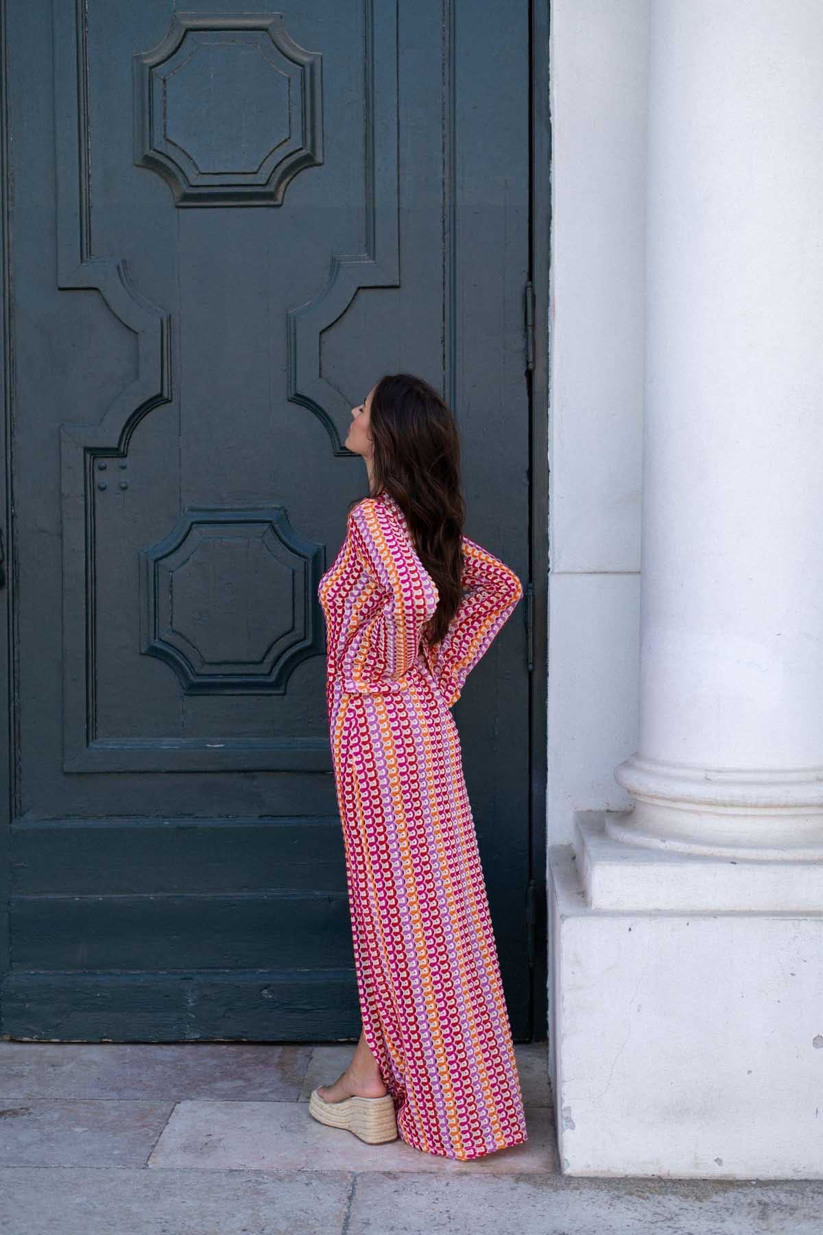Red - A woman models a textured, open-knit maxi dress featuring a deep V-neck, long bell sleeves, a front slit, and a vertical striped pattern, standing outdoors near pink flowers.