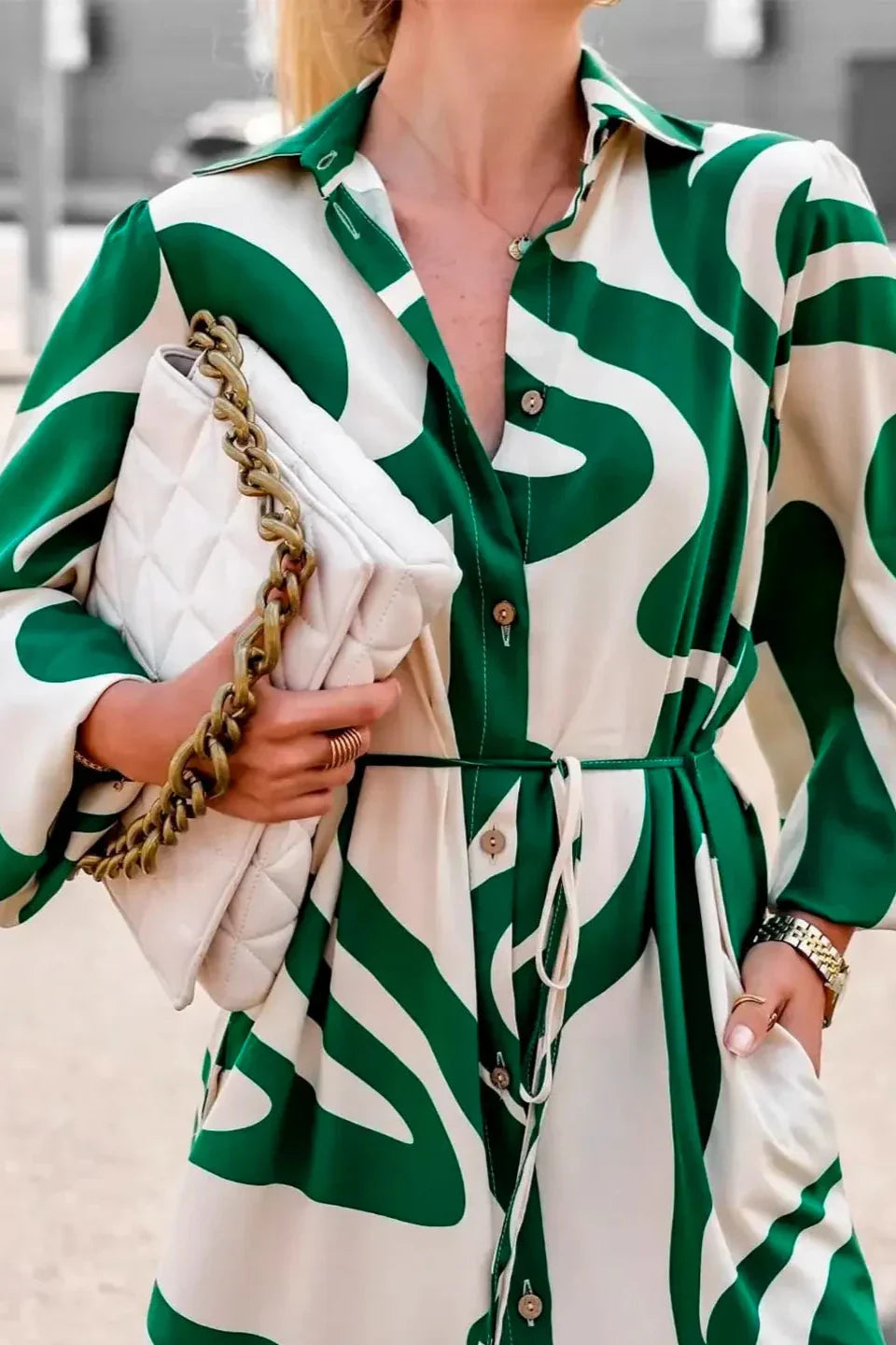 Green - A woman wearing a long, black and white abstract print shirt dress with a tie waist, blouson sleeves, and a high side slit, paired with white chunky loafers.