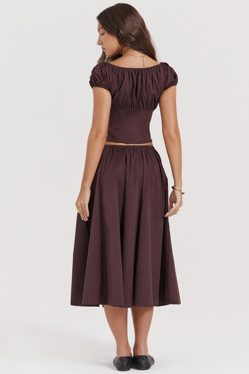 Brown - A woman models a dark brown midi dress featuring a ruched, button-front bodice with cap sleeves, daring waist cutouts with tie details, and a gathered flowing skirt, standing outdoors by rustic wooden doors.