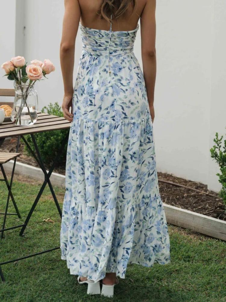 Blue - A woman wearing a white maxi dress with an orange and blue floral print, featuring a halter neckline, ruched bodice, and tiered skirt, standing outdoors.