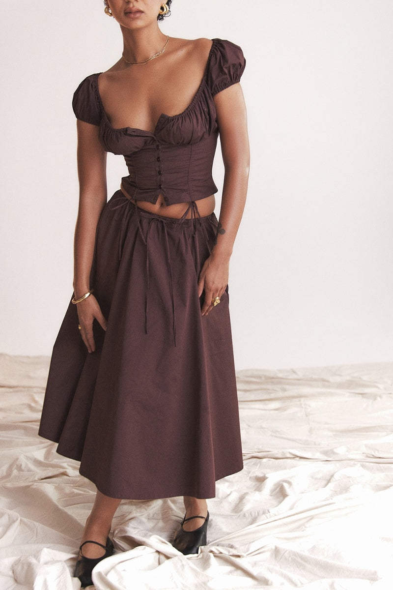 A woman models a dark brown midi dress featuring a ruched, button-front bodice with cap sleeves, daring waist cutouts with tie details, and a gathered flowing skirt, standing outdoors by rustic wooden doors.
