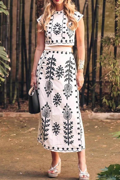 White - Women's two-piece set featuring a high-neck sleeveless crop top and a wrap maxi skirt with a side tie, adorned with contrasting folkloric embroidery, worn by a model standing on steps.