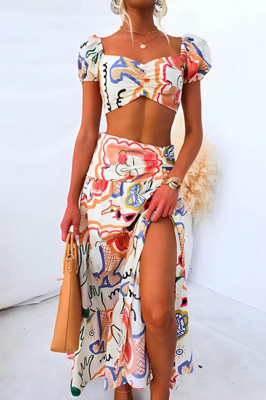 Women's two-piece maxi dress featuring a crop top with puff sleeves, a ruched bust, and a high-waisted skirt with a thigh-high slit, all in a colourful abstract graphic print.