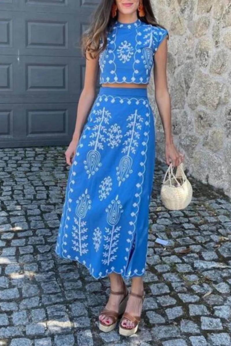 White - Women's two-piece set featuring a high-neck sleeveless crop top and a wrap maxi skirt with a side tie, adorned with contrasting folkloric embroidery, worn by a model standing on steps.