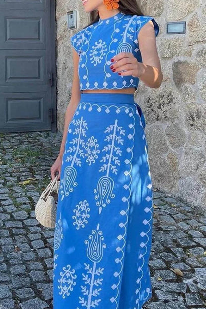 Blue - Women's two-piece set featuring a high-neck sleeveless crop top and a wrap maxi skirt with a side tie, adorned with contrasting folkloric embroidery, worn by a model standing on steps.