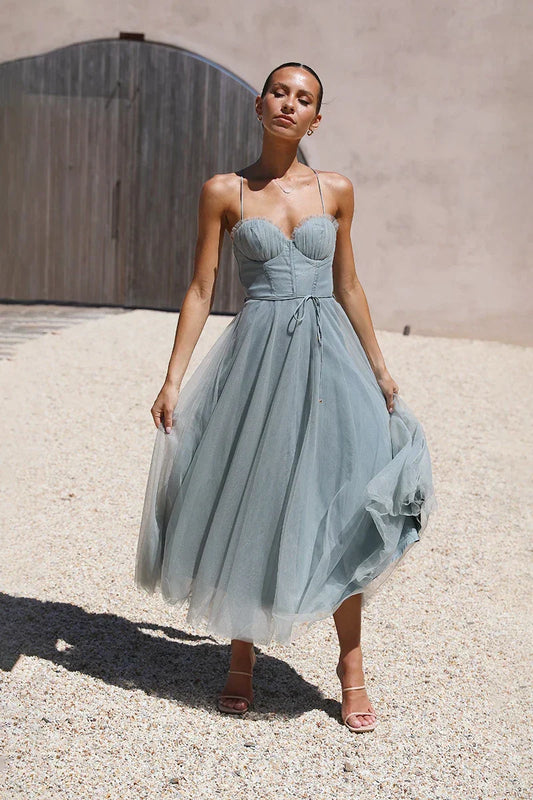 Light Blue - A woman modelling an elegant dusty blue midi dress featuring a structured corset bodice, thin straps, a tie waist, and a full A-line skirt with a sheer overlay, standing outdoors on a gravel path.