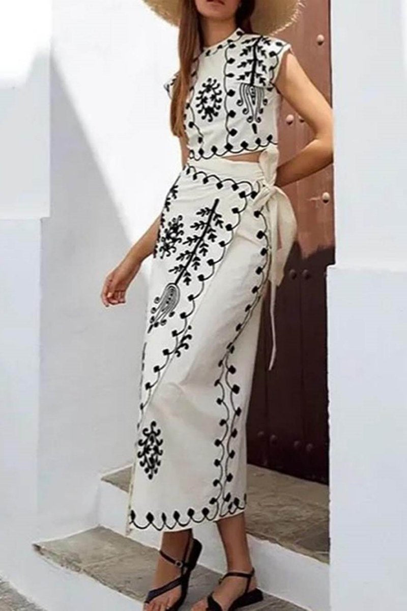 Women's two-piece set featuring a high-neck sleeveless crop top and a wrap maxi skirt with a side tie, adorned with contrasting folkloric embroidery, worn by a model standing on steps.