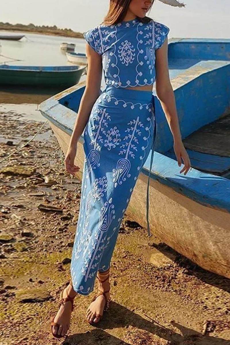 Blue - Women's two-piece set featuring a high-neck sleeveless crop top and a wrap maxi skirt with a side tie, adorned with contrasting folkloric embroidery, worn by a model standing on steps.