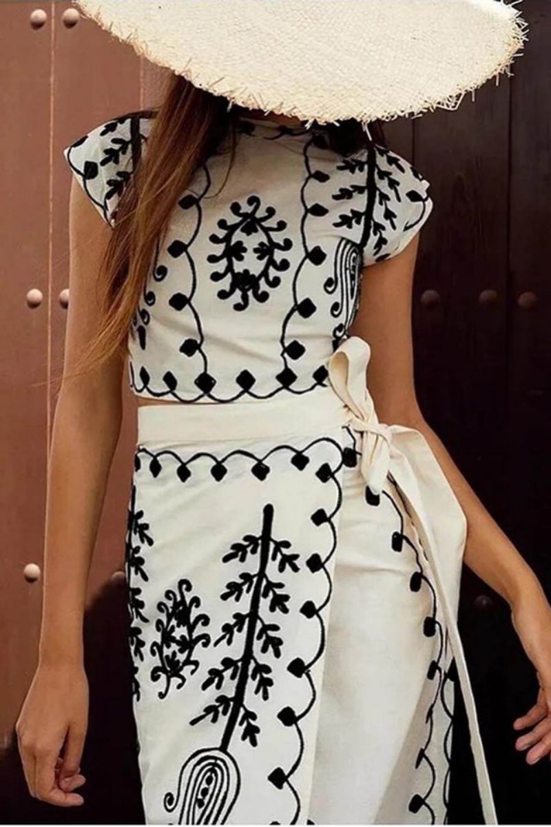 White - Women's two-piece set featuring a high-neck sleeveless crop top and a wrap maxi skirt with a side tie, adorned with contrasting folkloric embroidery, worn by a model standing on steps.
