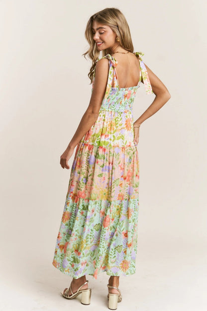 Multicolor - A woman wearing a vibrant floral print maxi dress featuring a smocked bodice, tie-shoulder straps, and a tiered skirt, paired with metallic heeled sandals.