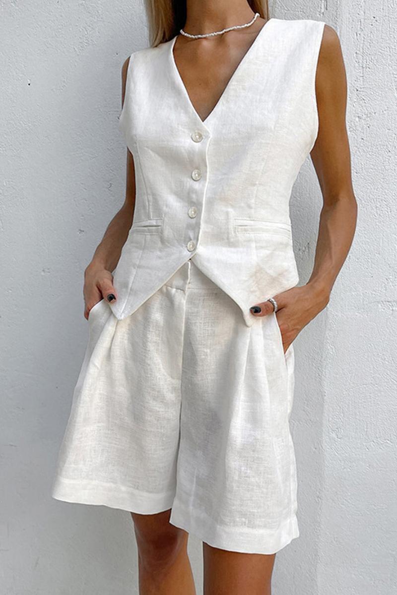 White - A woman wearing a white, sleeveless, button-front tailored waistcoat top paired with matching high-waisted pleated shorts, standing against a textured white wall.
