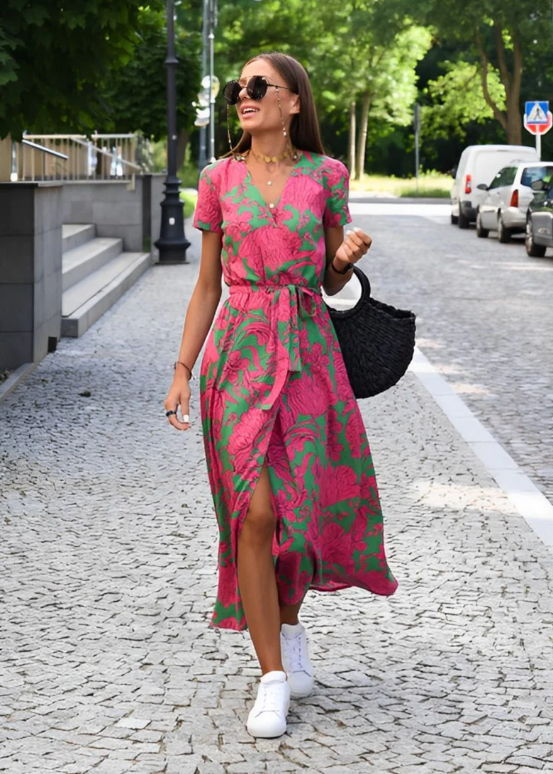 Green and Pink - A woman walking outdoors wearing a vibrant pink and green floral print midi dress with a V-neckline, short sleeves, a tie waist, and a front slit.