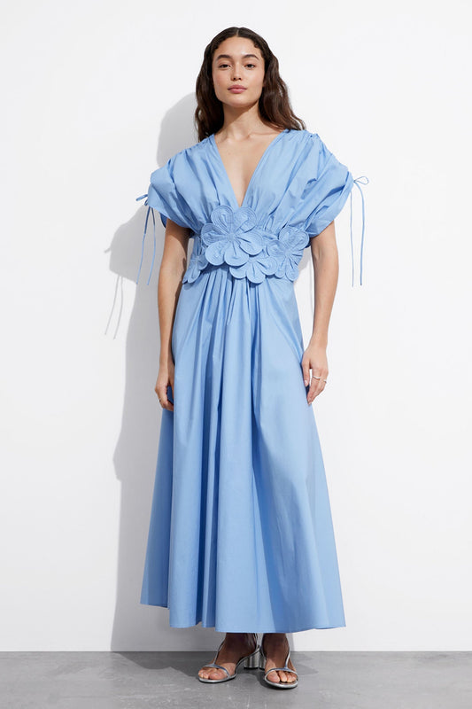 Blue - A woman wearing a light blue maxi dress featuring a deep V-neckline, tie-detail short sleeves, and large 3D floral appliqués at the waist.