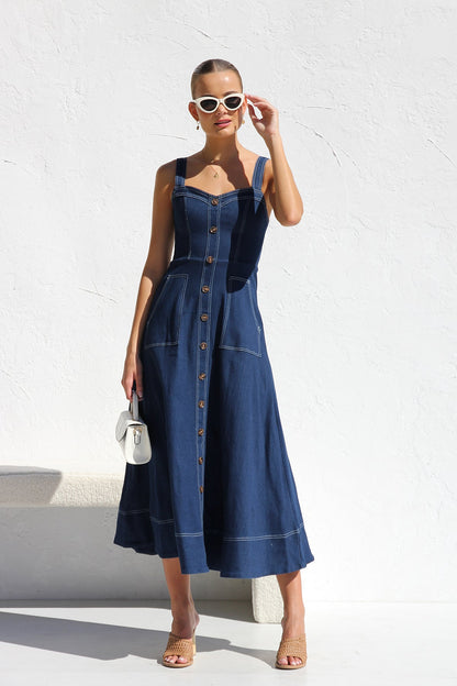 A woman models a dark blue, sleeveless A-line midi dress with contrast white topstitching and a full button-front. She wears white cat-eye sunglasses and holds a small white handbag, standing against a white textured wall.