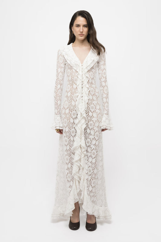 A woman wearing a sheer white floral lace maxi dress featuring a deep V-neckline, prominent cascading ruffles down the buttoned front, and long sleeves with flared ruffled cuffs.