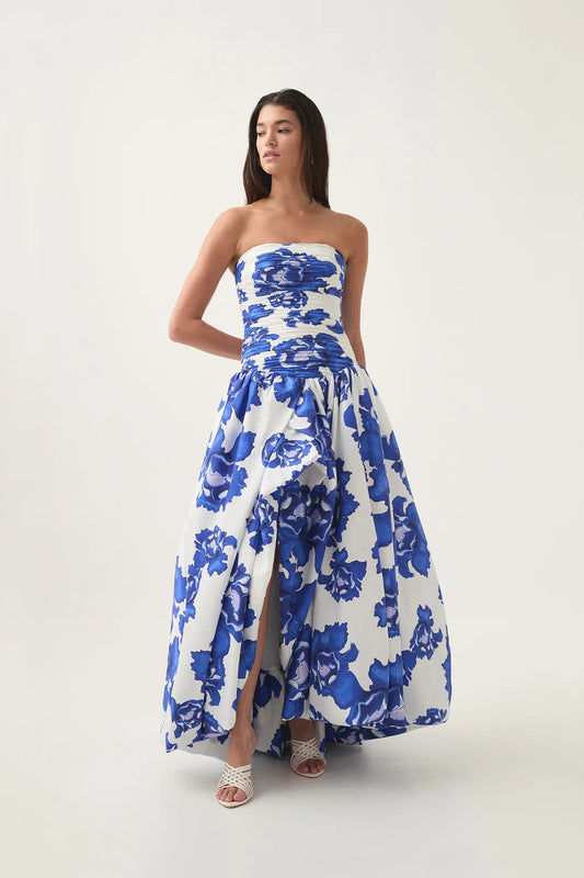 Boldly blooming cobalt - A woman models a strapless maxi dress featuring a white background with a large blue floral print. The dress has a ruched, fitted bodice, a dropped waist, and a full, voluminous skirt with a high front slit.