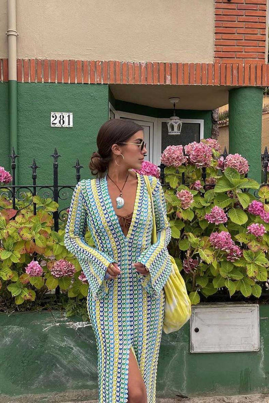Green - A woman models a textured, open-knit maxi dress featuring a deep V-neck, long bell sleeves, a front slit, and a vertical striped pattern, standing outdoors near pink flowers.