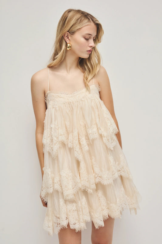 A woman models a cream-coloured, tiered mini dress featuring semi-sheer ruffles, delicate lace trim, and thin spaghetti straps.