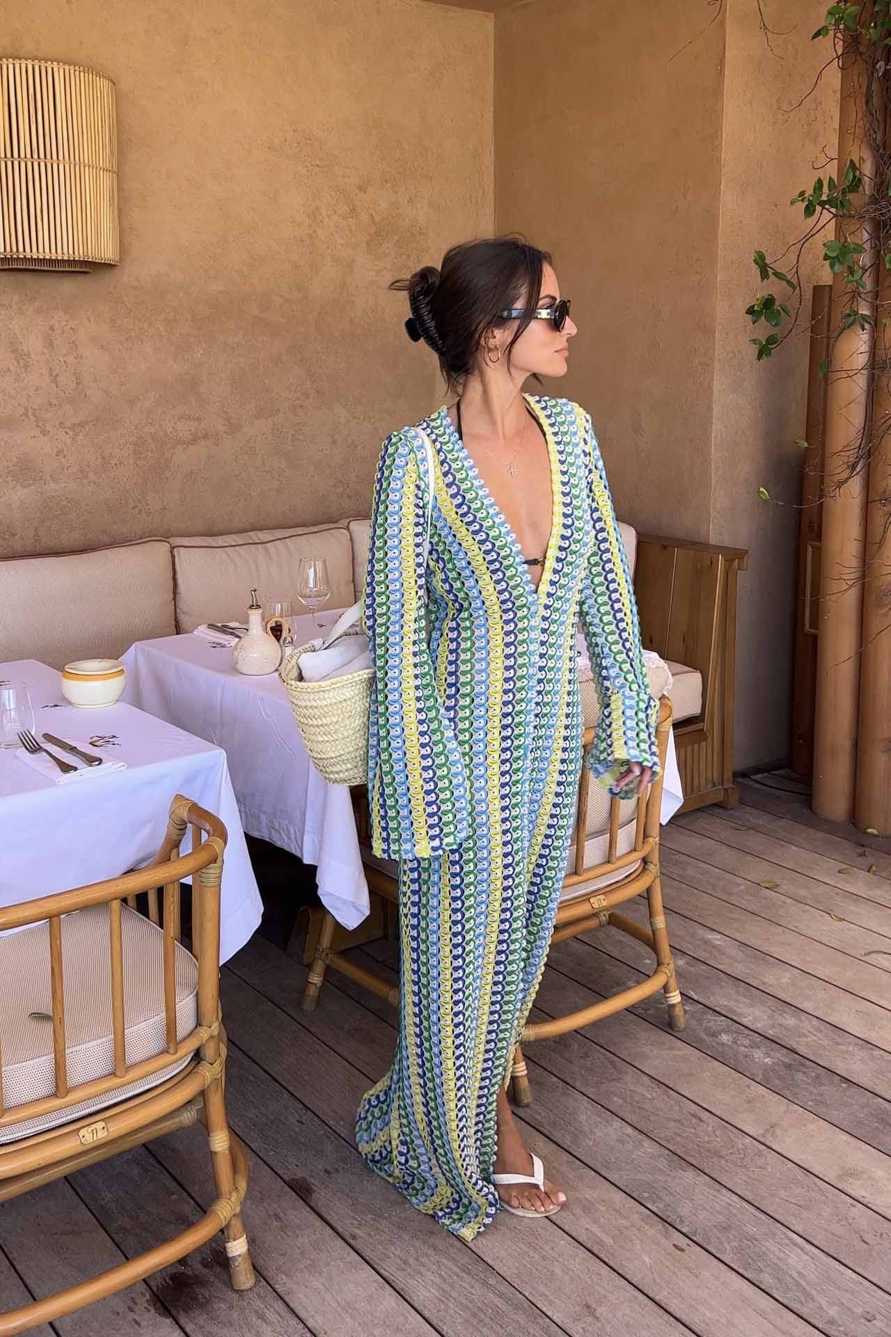 Green - A woman models a textured, open-knit maxi dress featuring a deep V-neck, long bell sleeves, a front slit, and a vertical striped pattern, standing outdoors near pink flowers.