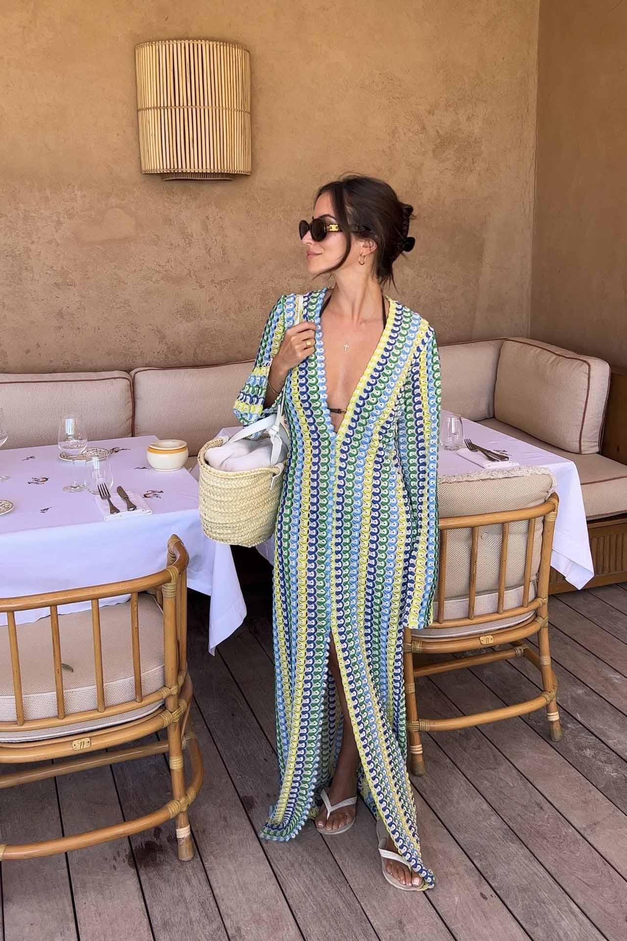 Green - A woman models a textured, open-knit maxi dress featuring a deep V-neck, long bell sleeves, a front slit, and a vertical striped pattern, standing outdoors near pink flowers.