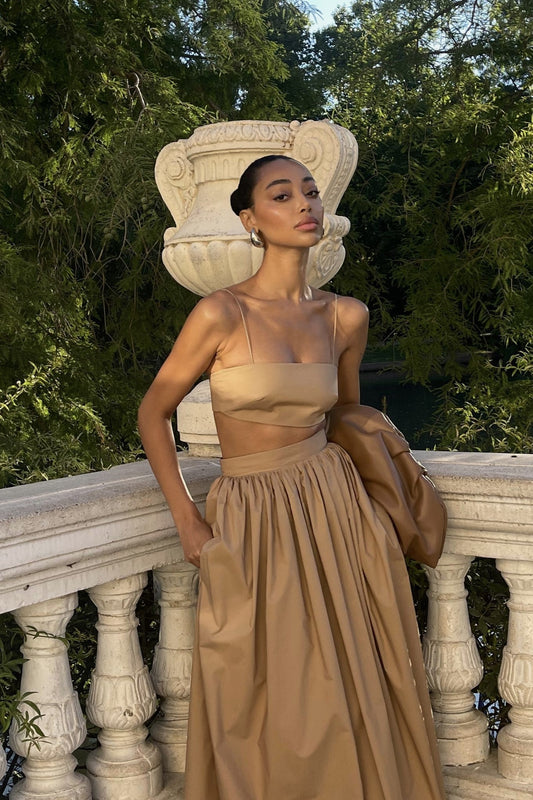 A fashion model wearing a chic, solid-coloured two-piece maxi dress featuring a minimalist spaghetti strap crop top and a high-waisted, voluminous gathered skirt with side pockets, posing outdoors against a classic stone balustrade.