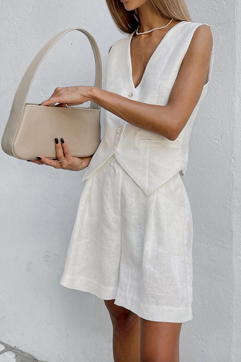 White - A woman wearing a white, sleeveless, button-front tailored waistcoat top paired with matching high-waisted pleated shorts, standing against a textured white wall.