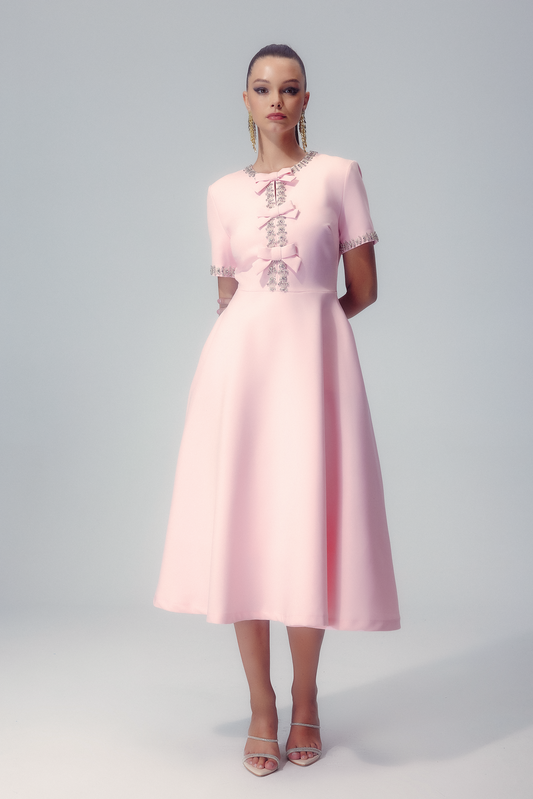 Pink - A woman modelling a light pink, short-sleeved midi dress featuring an A-line silhouette, three front bows, and sparkling crystal embellishments along the neckline, cuffs, and centre bodice.