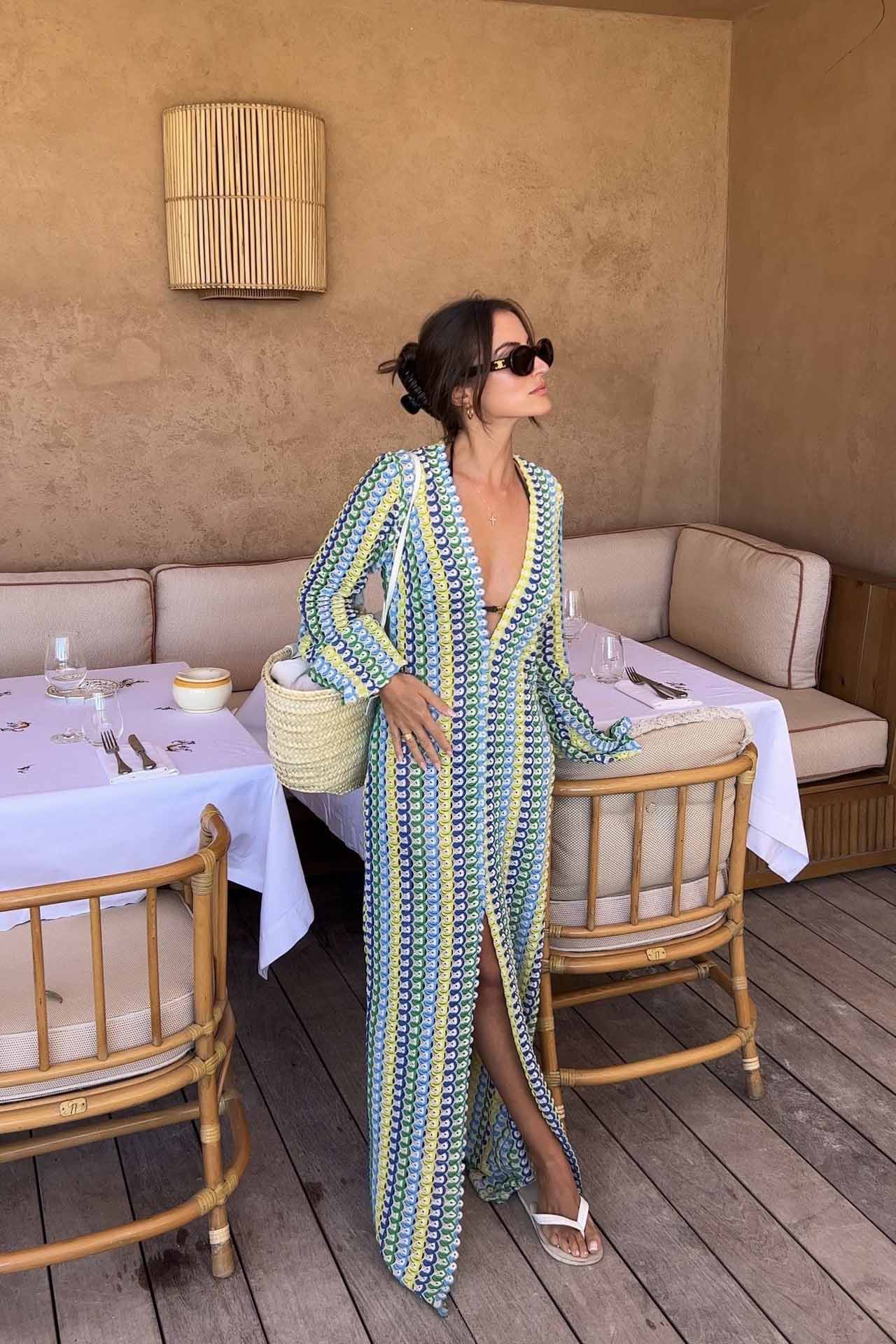 Green - A woman models a textured, open-knit maxi dress featuring a deep V-neck, long bell sleeves, a front slit, and a vertical striped pattern, standing outdoors near pink flowers.