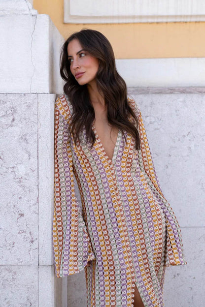 Green - A woman models a textured, open-knit maxi dress featuring a deep V-neck, long bell sleeves, a front slit, and a vertical striped pattern, standing outdoors near pink flowers.