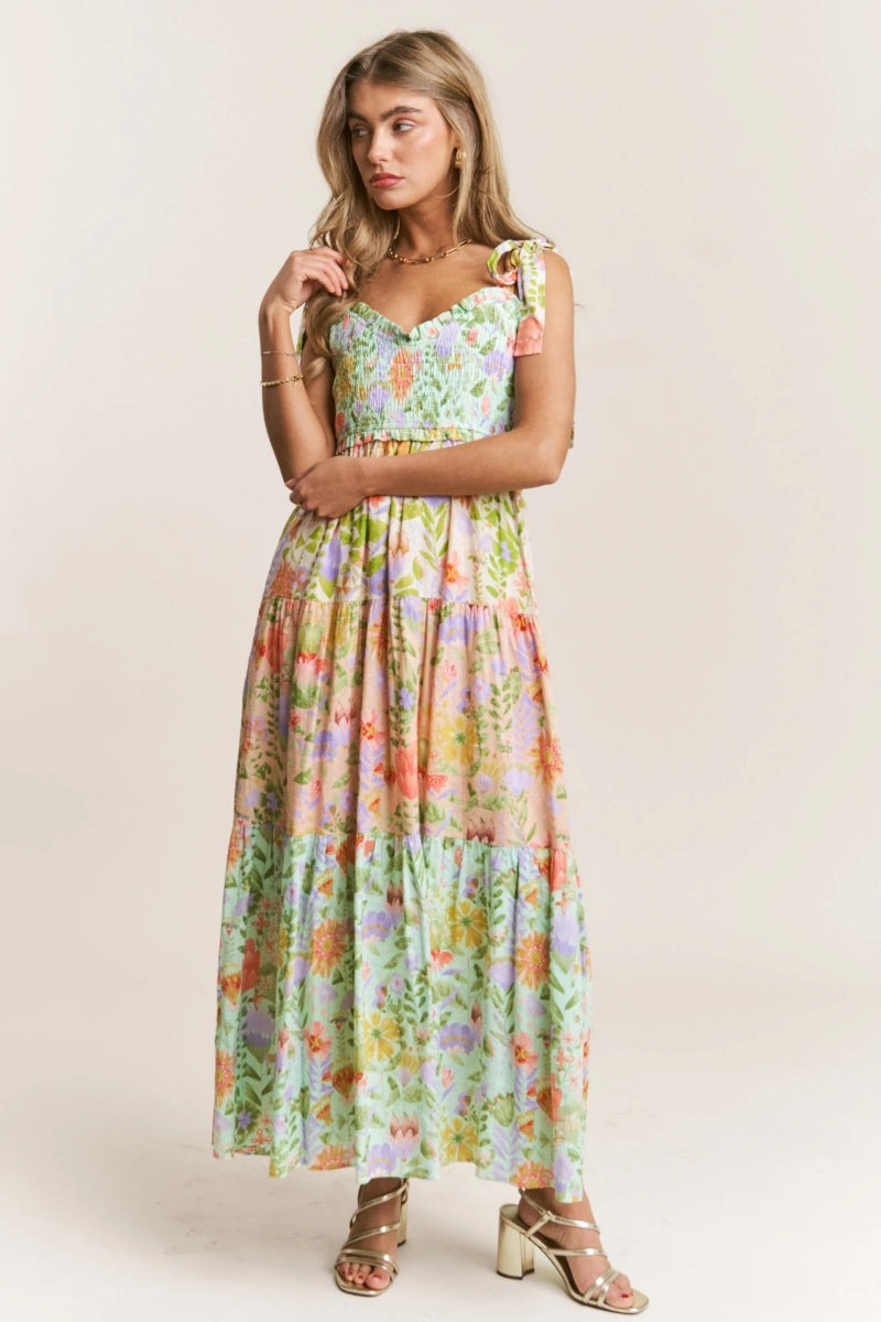 A woman wearing a vibrant floral print maxi dress featuring a smocked bodice, tie-shoulder straps, and a tiered skirt, paired with metallic heeled sandals.