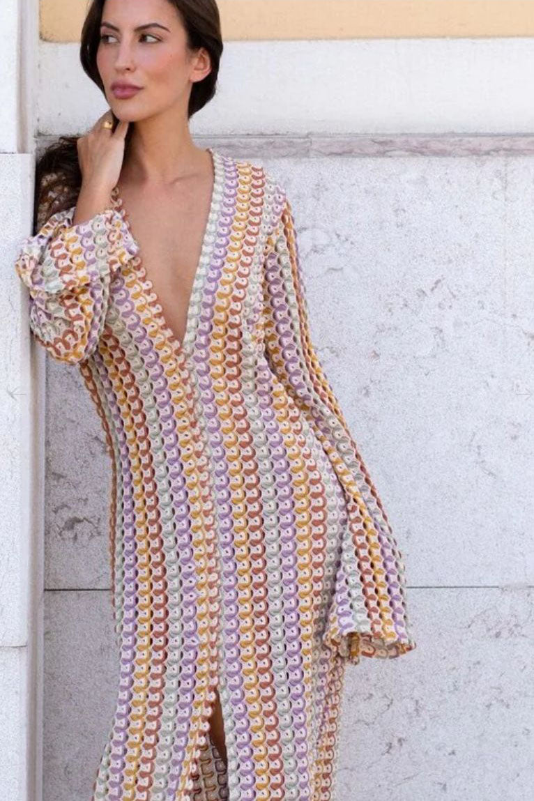 Yellow - A woman models a textured, open-knit maxi dress featuring a deep V-neck, long bell sleeves, a front slit, and a vertical striped pattern, standing outdoors near pink flowers.