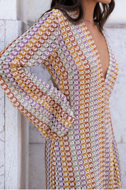 Yellow - A woman models a textured, open-knit maxi dress featuring a deep V-neck, long bell sleeves, a front slit, and a vertical striped pattern, standing outdoors near pink flowers.