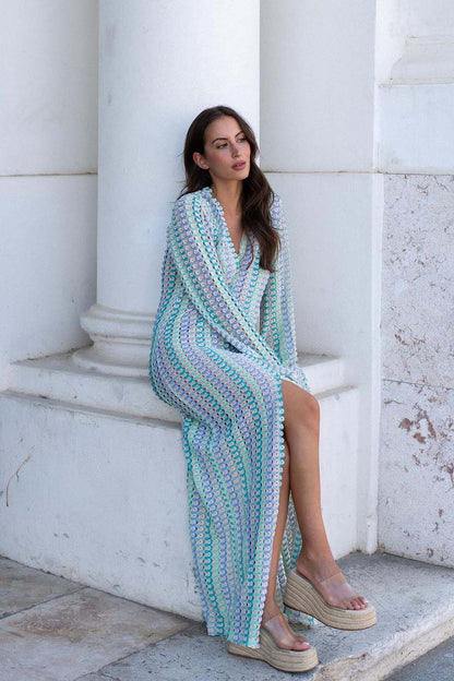 Blue - A woman models a textured, open-knit maxi dress featuring a deep V-neck, long bell sleeves, a front slit, and a vertical striped pattern, standing outdoors near pink flowers.