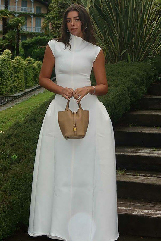 Woman wearing an elegant white sleeveless maxi dress with a high neckline and a structured, voluminous skirt, standing on outdoor brick steps at night.