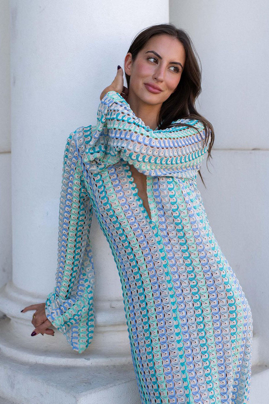 Blue - A woman models a textured, open-knit maxi dress featuring a deep V-neck, long bell sleeves, a front slit, and a vertical striped pattern, standing outdoors near pink flowers.