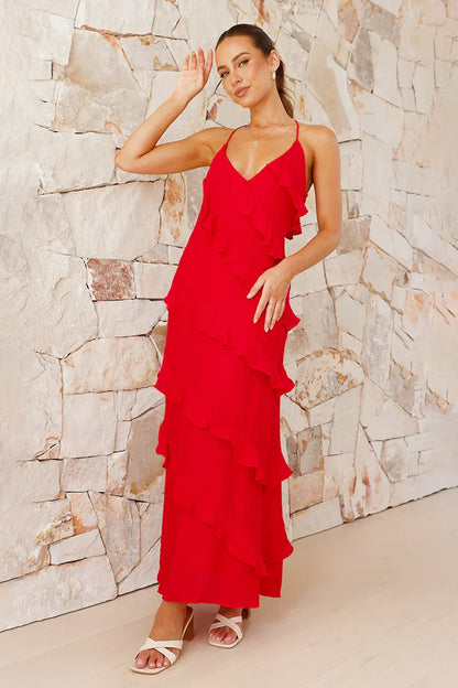 A woman modelling a vibrant red, floor-length maxi dress featuring a V-neckline, thin straps, and elegant asymmetrical cascading ruffles down the front, standing against a light textured stone wall.