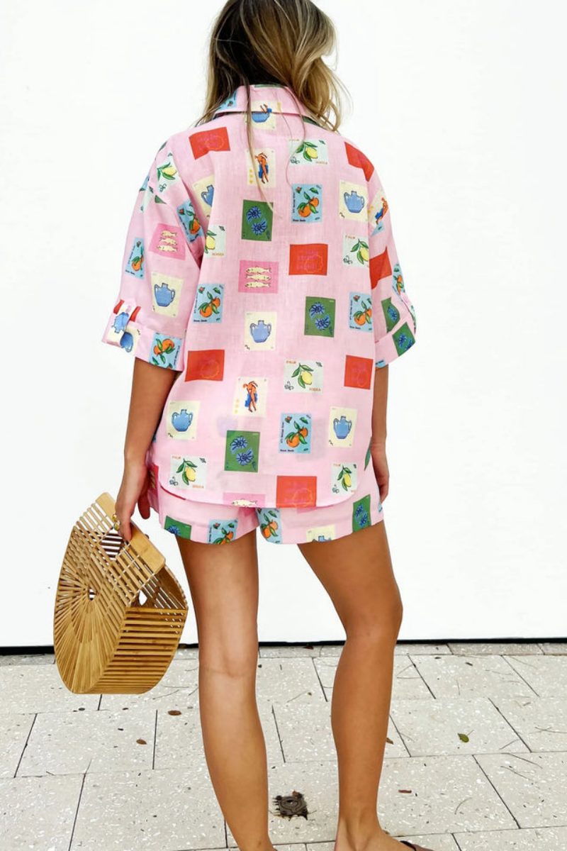 Pink - Women's pink short-sleeve belted playsuit featuring a colourful Mediterranean novelty print with oranges and vases, styled with sunglasses and a bamboo bag.