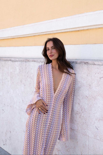 Pink - A woman models a textured, open-knit maxi dress featuring a deep V-neck, long bell sleeves, a front slit, and a vertical striped pattern, standing outdoors near pink flowers.