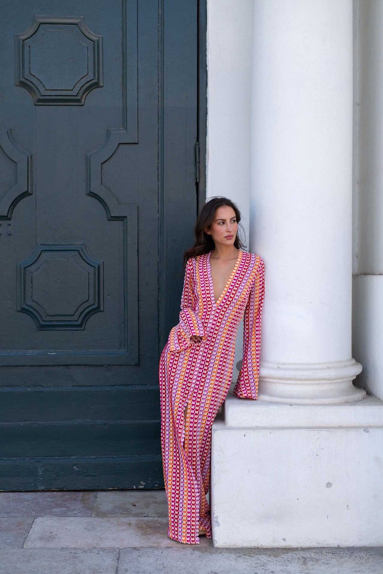 Red - A woman models a textured, open-knit maxi dress featuring a deep V-neck, long bell sleeves, a front slit, and a vertical striped pattern, standing outdoors near pink flowers.