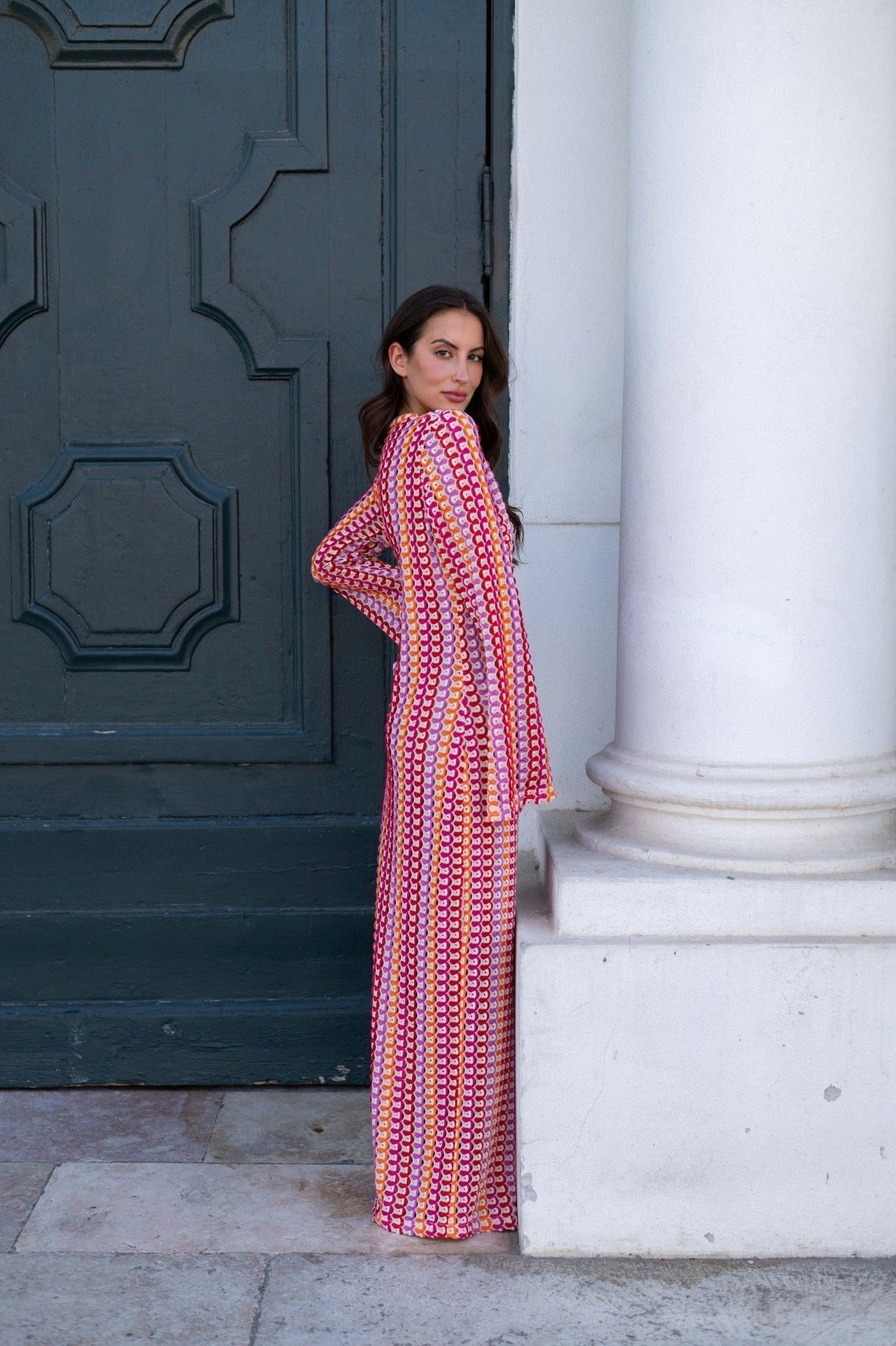 Red - A woman models a textured, open-knit maxi dress featuring a deep V-neck, long bell sleeves, a front slit, and a vertical striped pattern, standing outdoors near pink flowers.