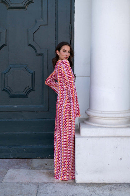 Red - A woman models a textured, open-knit maxi dress featuring a deep V-neck, long bell sleeves, a front slit, and a vertical striped pattern, standing outdoors near pink flowers.