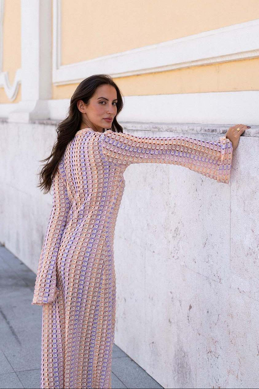 Pink - A woman models a textured, open-knit maxi dress featuring a deep V-neck, long bell sleeves, a front slit, and a vertical striped pattern, standing outdoors near pink flowers.