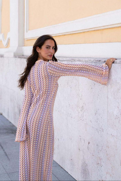 Pink - A woman models a textured, open-knit maxi dress featuring a deep V-neck, long bell sleeves, a front slit, and a vertical striped pattern, standing outdoors near pink flowers.