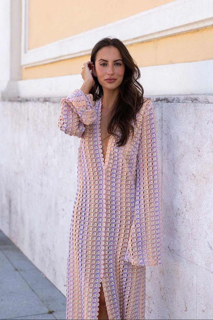 Pink - A woman models a textured, open-knit maxi dress featuring a deep V-neck, long bell sleeves, a front slit, and a vertical striped pattern, standing outdoors near pink flowers.