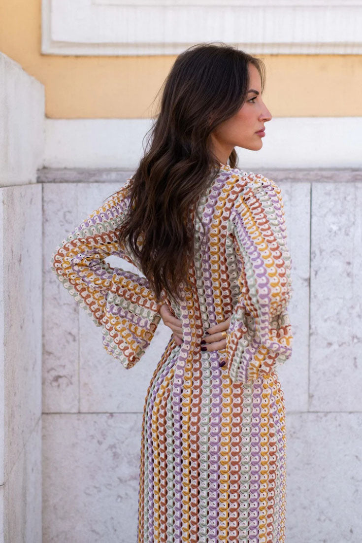 Yellow - A woman models a textured, open-knit maxi dress featuring a deep V-neck, long bell sleeves, a front slit, and a vertical striped pattern, standing outdoors near pink flowers.