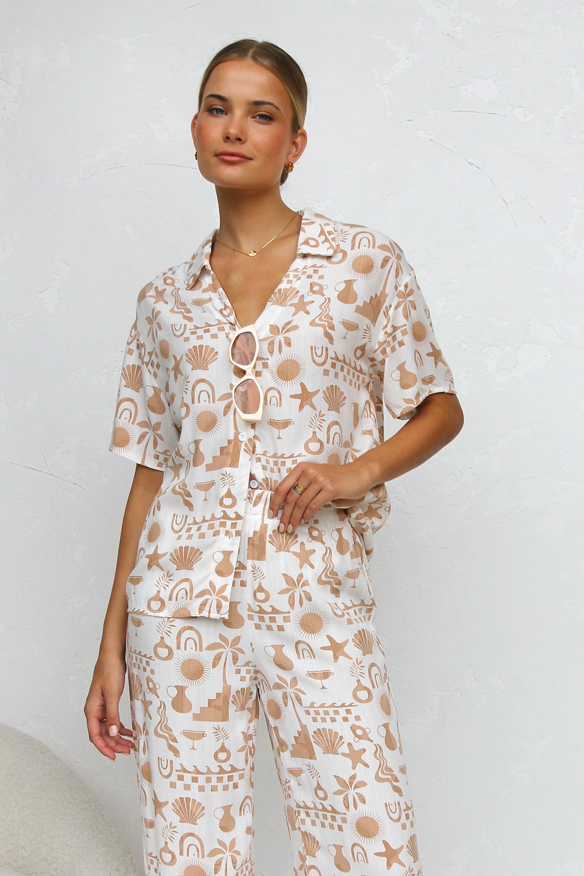 A woman modelling a relaxed-fit, wide-leg trouser and matching short-sleeve shirt co-ord set, featuring a beige coastal vacation print on a white background.
