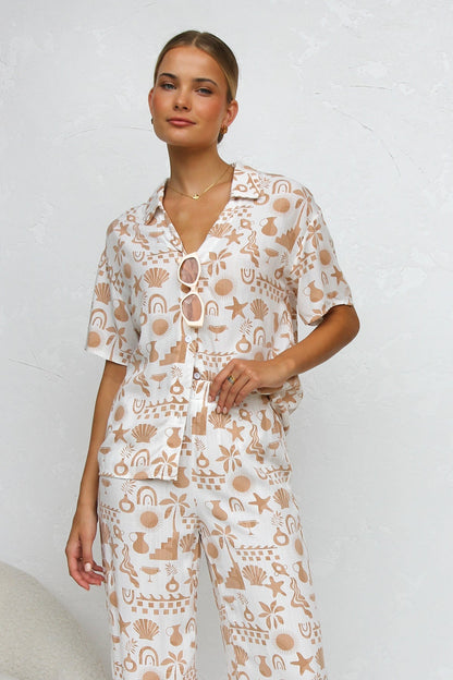 A woman modelling a relaxed-fit, wide-leg trouser and matching short-sleeve shirt co-ord set, featuring a beige coastal vacation print on a white background.