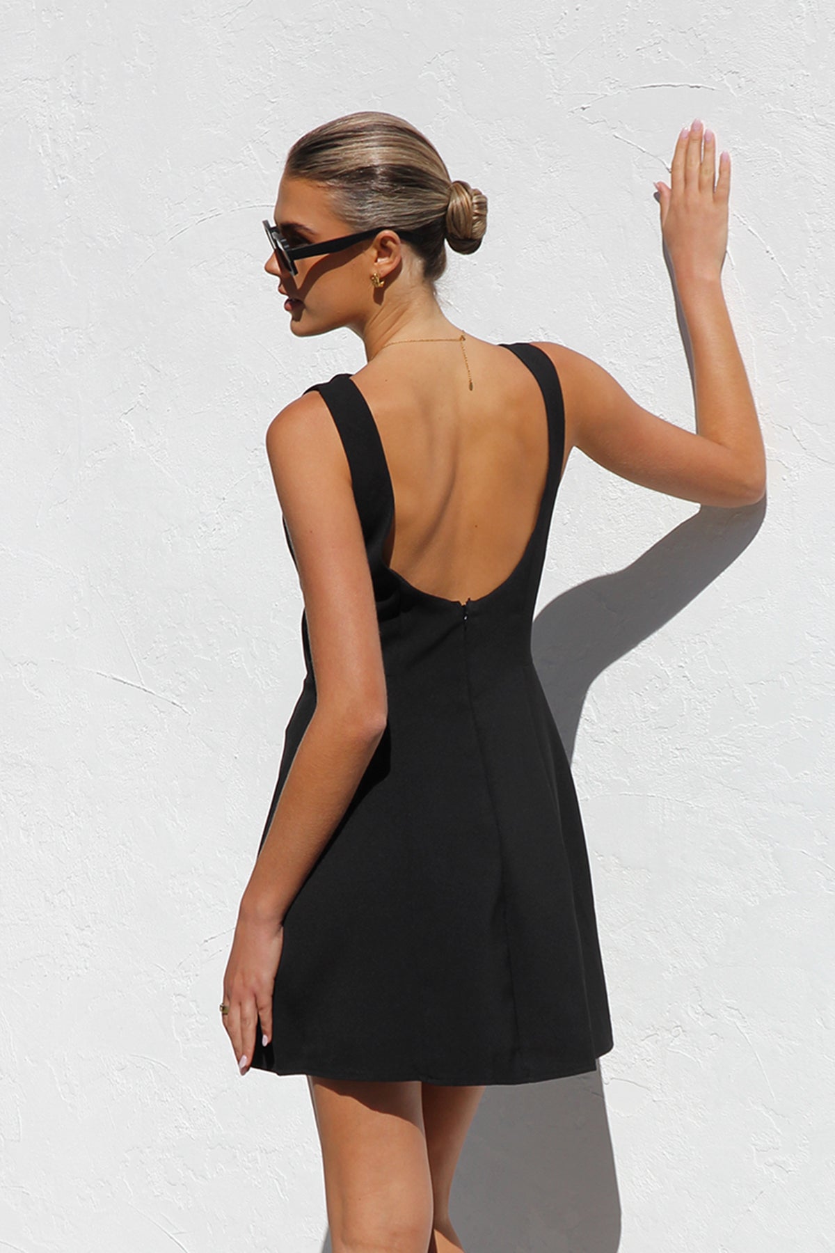 Black - A woman with blonde hair in a bun, wearing black rectangular sunglasses and a sleeveless, black, A-line mini dress with bodice seams, stands against a white textured wall in bright sunlight.