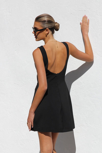 Black - A woman with blonde hair in a bun, wearing black rectangular sunglasses and a sleeveless, black, A-line mini dress with bodice seams, stands against a white textured wall in bright sunlight.