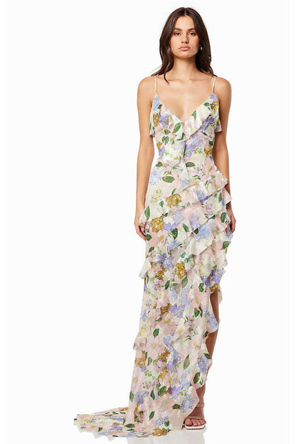 Green - A woman wearing a floor-length, ruffled maxi dress with a pastel floral print on a sheer cream background, featuring spaghetti straps and an asymmetric hem.